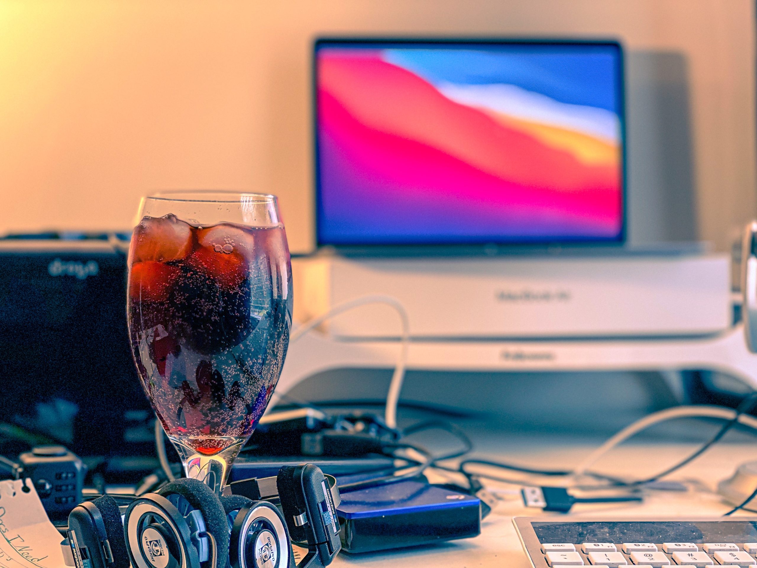 Glass goblet full of maroon bubbly liquid and ice in front of a laptop computer on a stand and other computer equipment