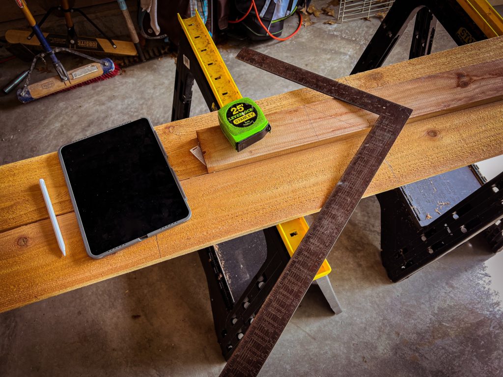 Some pieces of wood sit on sawhorses. On the wood sits an iPad and Apple Pencil, a neon green Stanley tape measure and a carpenter’s square.