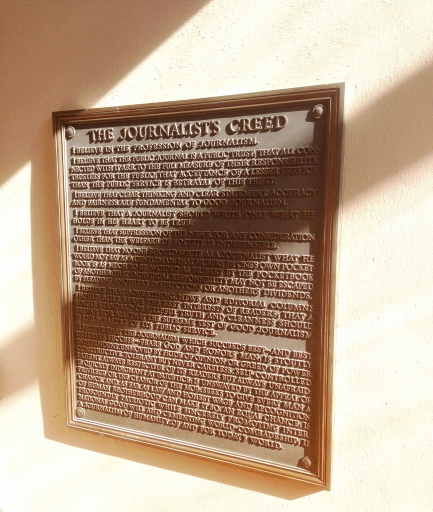 A bronze plaque with raised letters. It displays "The Journalist's Creed" in full. It is hanging on a  wall, shot at an angle, with sunlight and sharp shadows falling across the wall and the plaque.