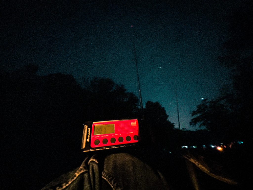 A bright red radio with 90.7 visible on the digital screen in front of a night scene, trees silhouetted in front of a dark sky with a few stars. 