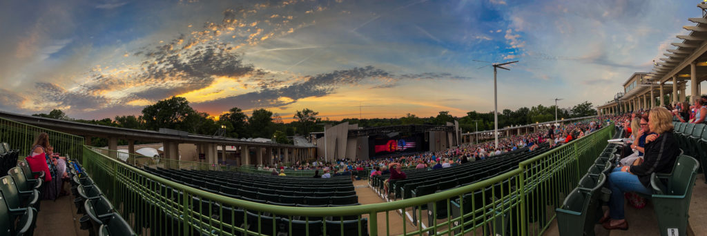 A panorama of an outdoor theater:  a blue sky with clouds, it is orange at the horizon, though the sun is not visible. The foreground is a sea of green theater seats, leading down very far to a stage. An image on a curtain or screen on the stage shows two closed fists meeting knuckles-to-knuckles. The left fist is red with a gold hammer and sickle. The right fist has the Stars and Stripes of the American flag. A tall overhead fan on a post rises from the seats on the right.