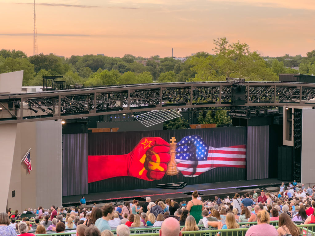 A theater stage, with people's heads in the foreground and an orange sky in the background. An image on a curtain or screen on the stage shows two closed fists meeting knuckles-to-knuckles. The left fist is red with a gold hammer and sickle. The right fist has the Stars and Stripes of the American flag. A tall overhead fan on a post rises from the seats on the right. The corner of a large chess board can be seen peeking out from below the curtain in the center of the stage. An American flag hangs to the left of the stage (from the viewer's perspective).